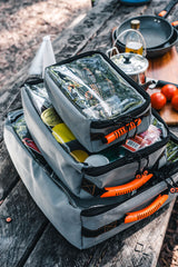 Stacked Wildtrak canvas bags for camping on a picnic table with outdoor cooking gear.